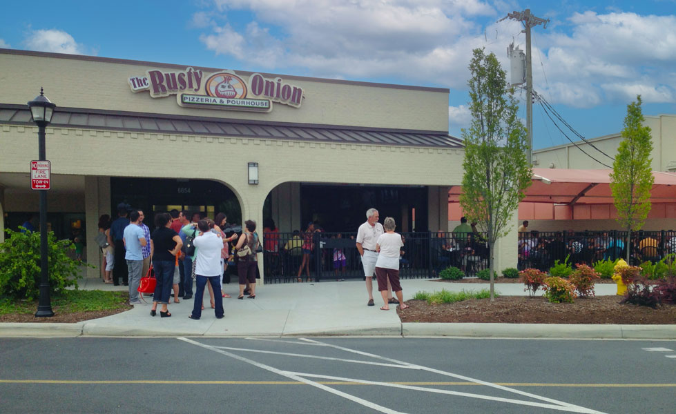 A retail space in a strip mall with a beige exterior and a line of people in front with an led sign on the facade that reads The Rusty Onion Pizzeria and Pourhouse